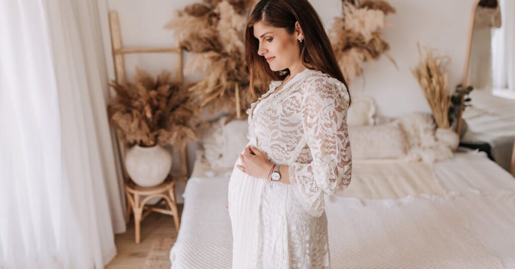 A pregnant woman in a white lace dress stands in a bohemian-style bedroom, embracing maternity.