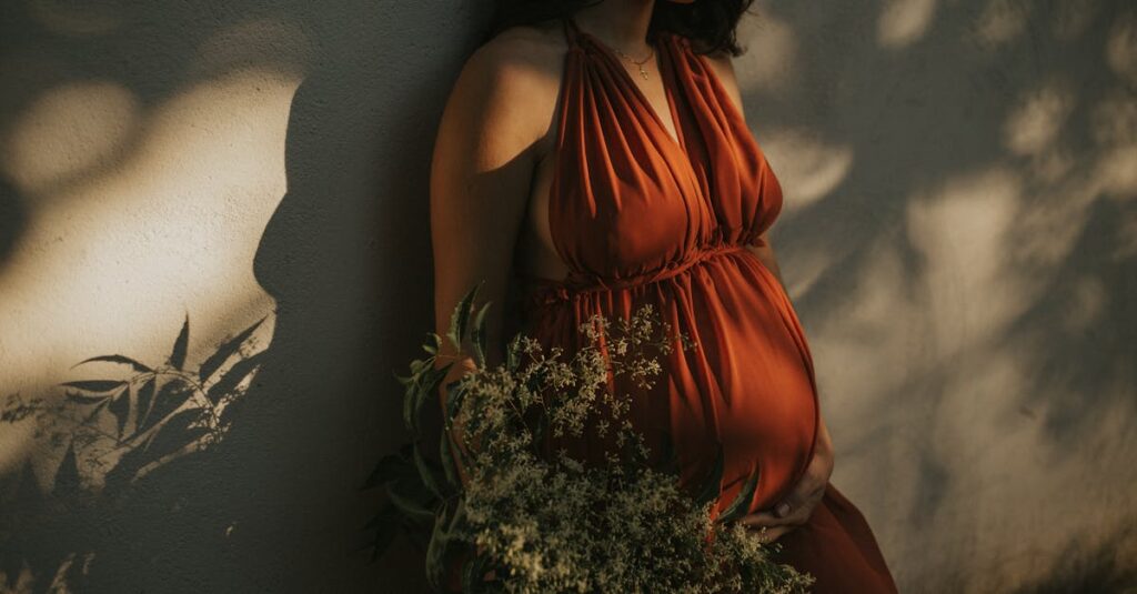 A serene pregnant woman in a vibrant dress holds flowers against a textured wall.