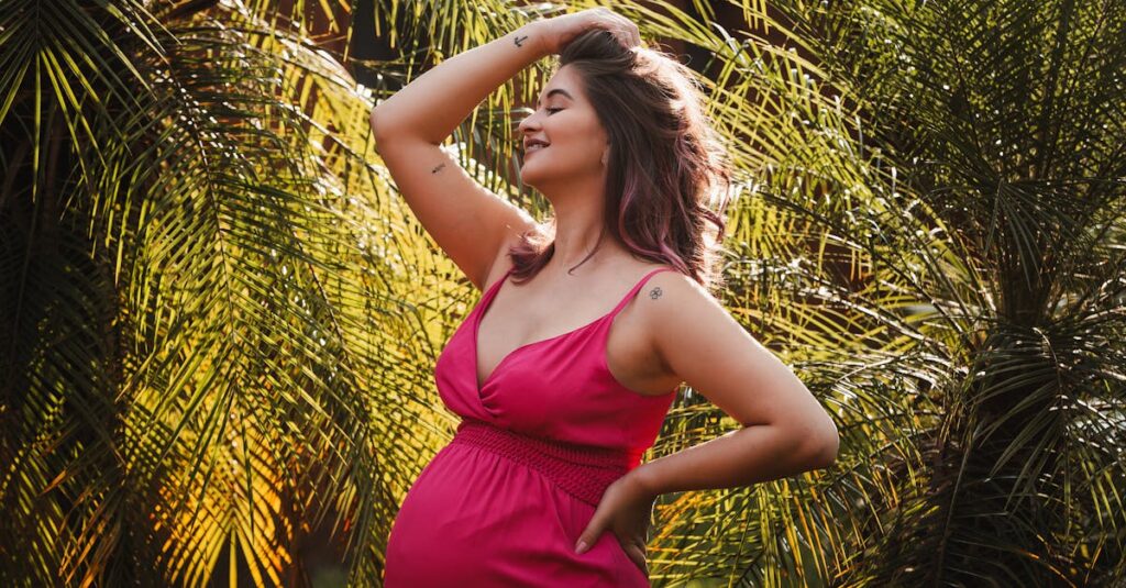 A pregnant woman in a vibrant pink dress poses happily among lush green plants outdoors.