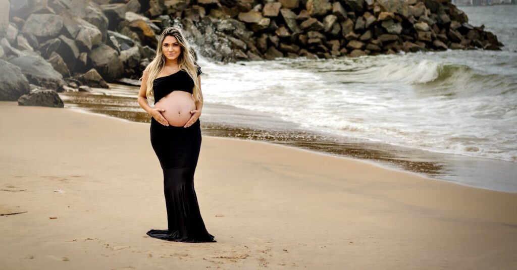 Pregnant woman in black dress standing on a sandy beach with rocky background, embracing motherhood.