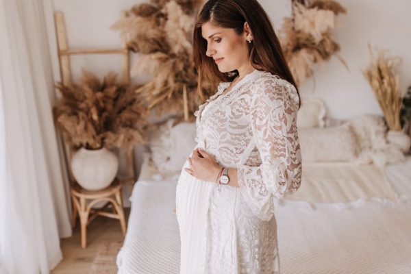 A pregnant woman in a white lace dress stands in a bohemian-style bedroom, embracing maternity.